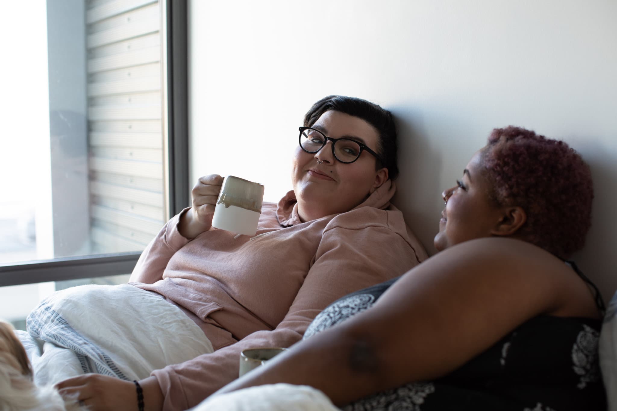 Two women laying in bed drinking coffee looking at each other